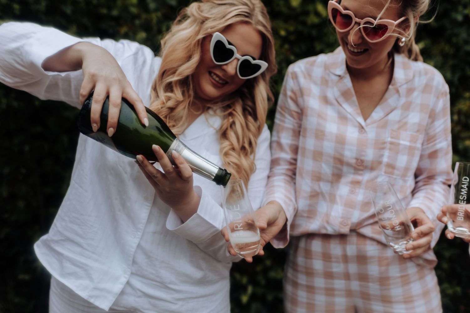 Two people in white and checkered outfits pour champagne into glasses outdoors.
