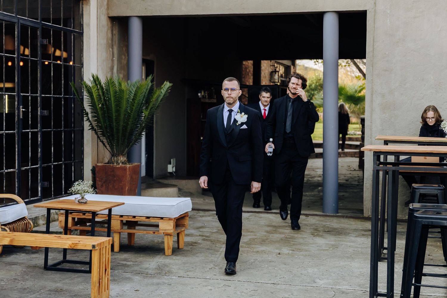People in formal attire walking through outdoor patio area with wooden benches and potted plants at venue entrance.