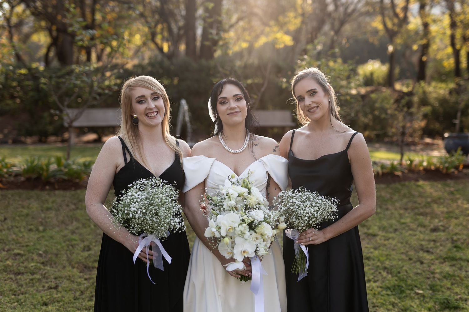 Three women in black and white dresses pose together holding wedding bouquets in a sunlit outdoor setting.