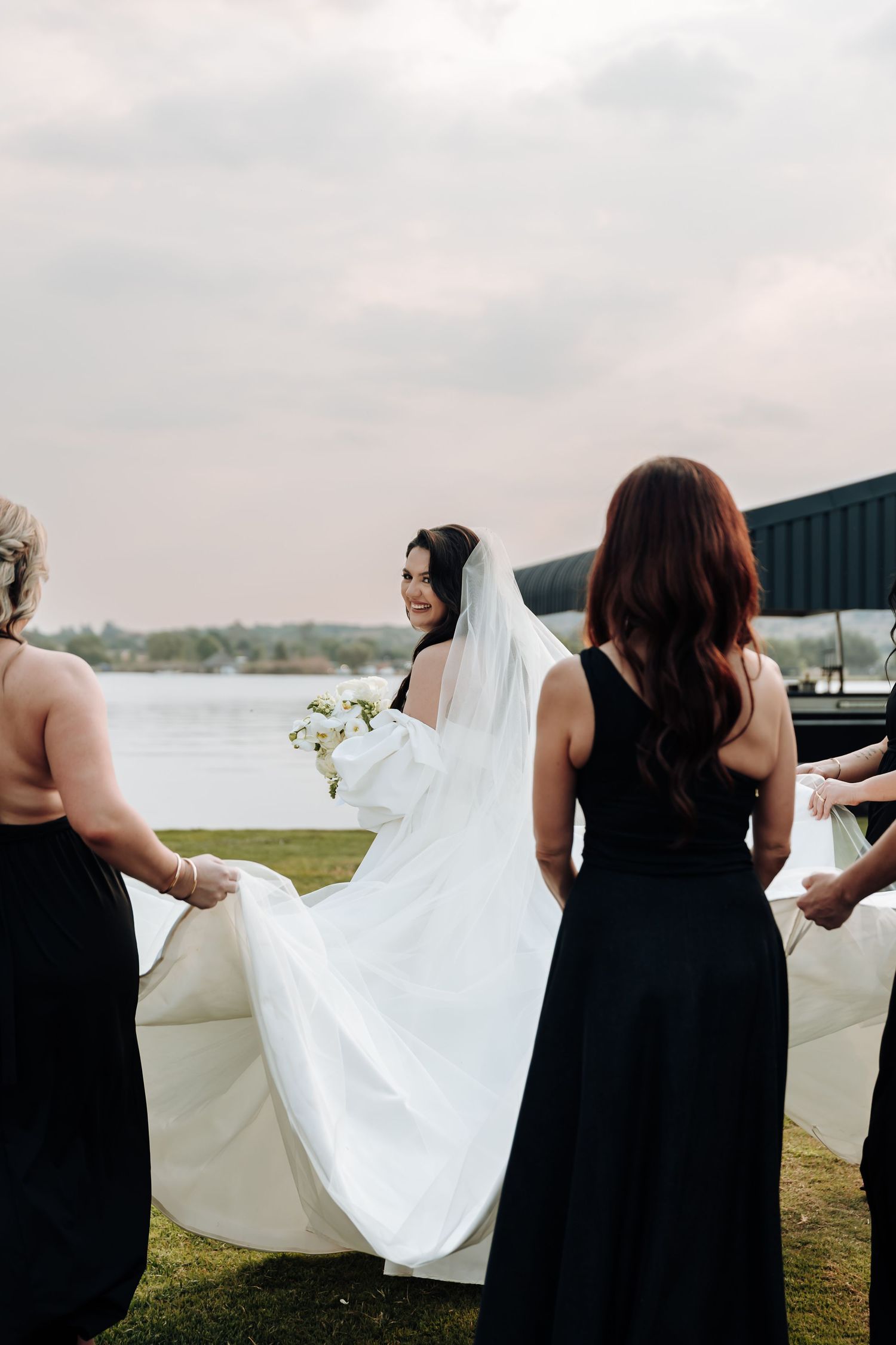 Bridesmaids in black dresses help adjust a flowing white wedding dress train on a grassy waterfront setting.
