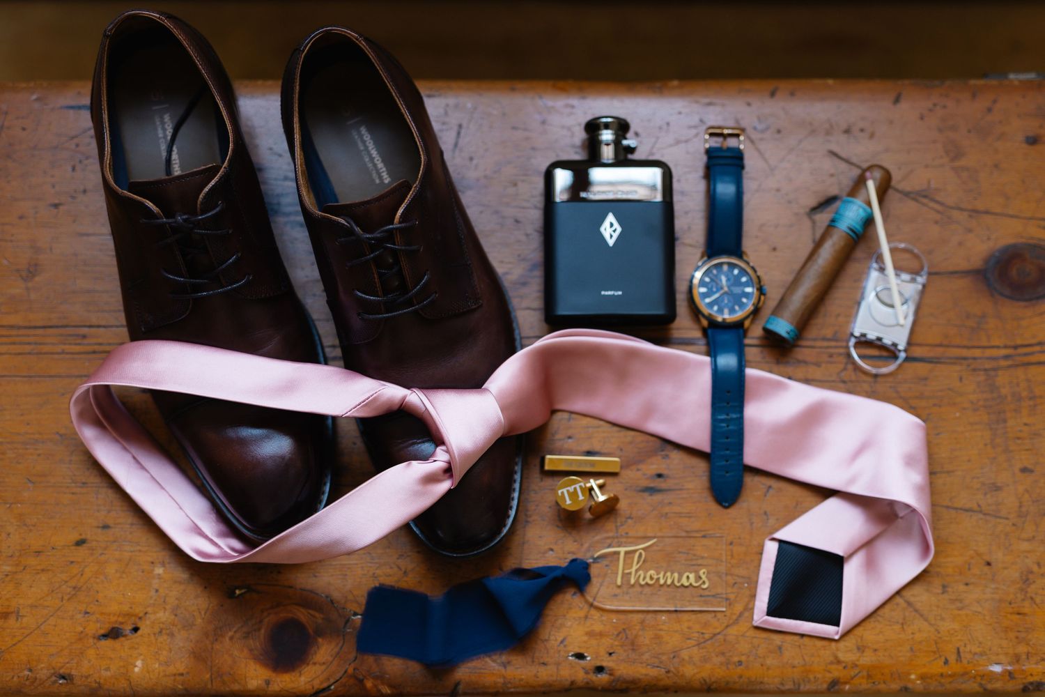 Pink bowtie and formal accessories laid out on rustic wooden table with black shoes and watch.