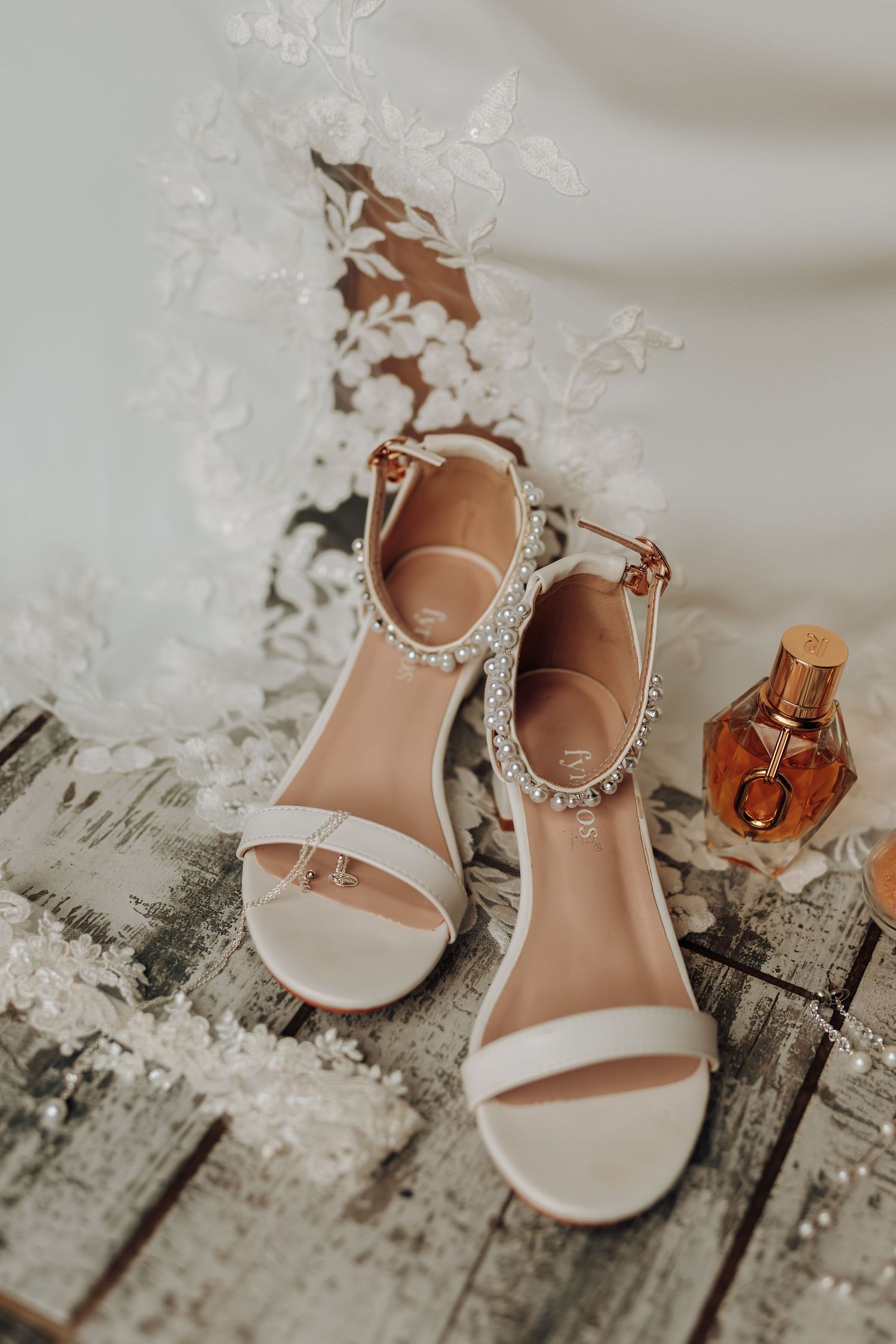 White bridal heels with crystal embellishments sit elegantly on a rustic wooden surface beside a perfume bottle.