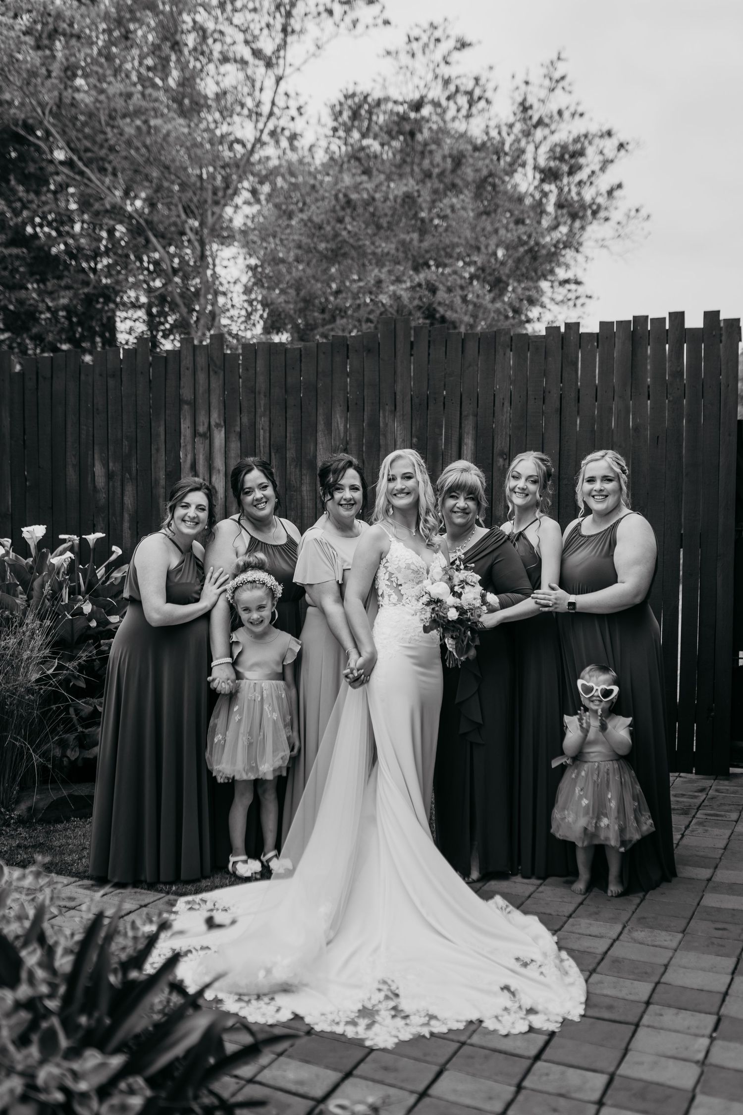 Black and white wedding photo showing a bridal party in dark gowns and flower girls posing against a wooden fence.