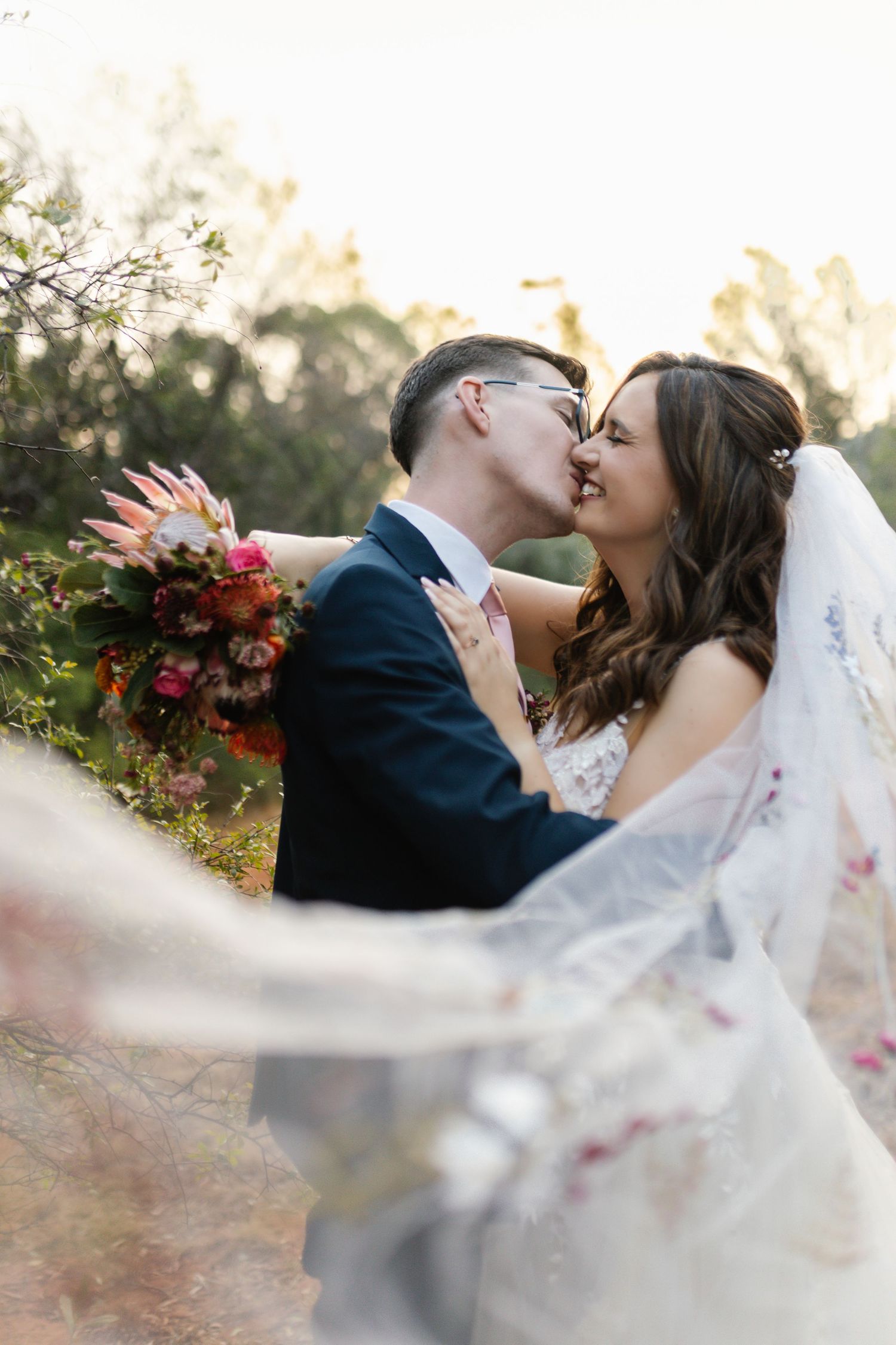 Newlyweds share romantic kiss outdoors with flowing veil and vibrant autumn bouquet at sunset.