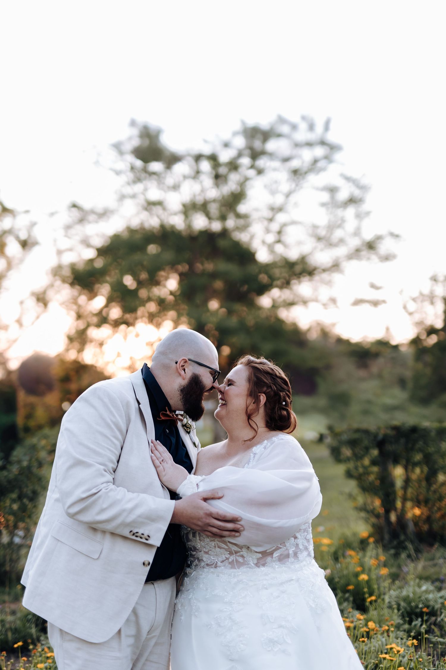 A romantic couple shares tender moments in a field at sunset wearing white formal attire.