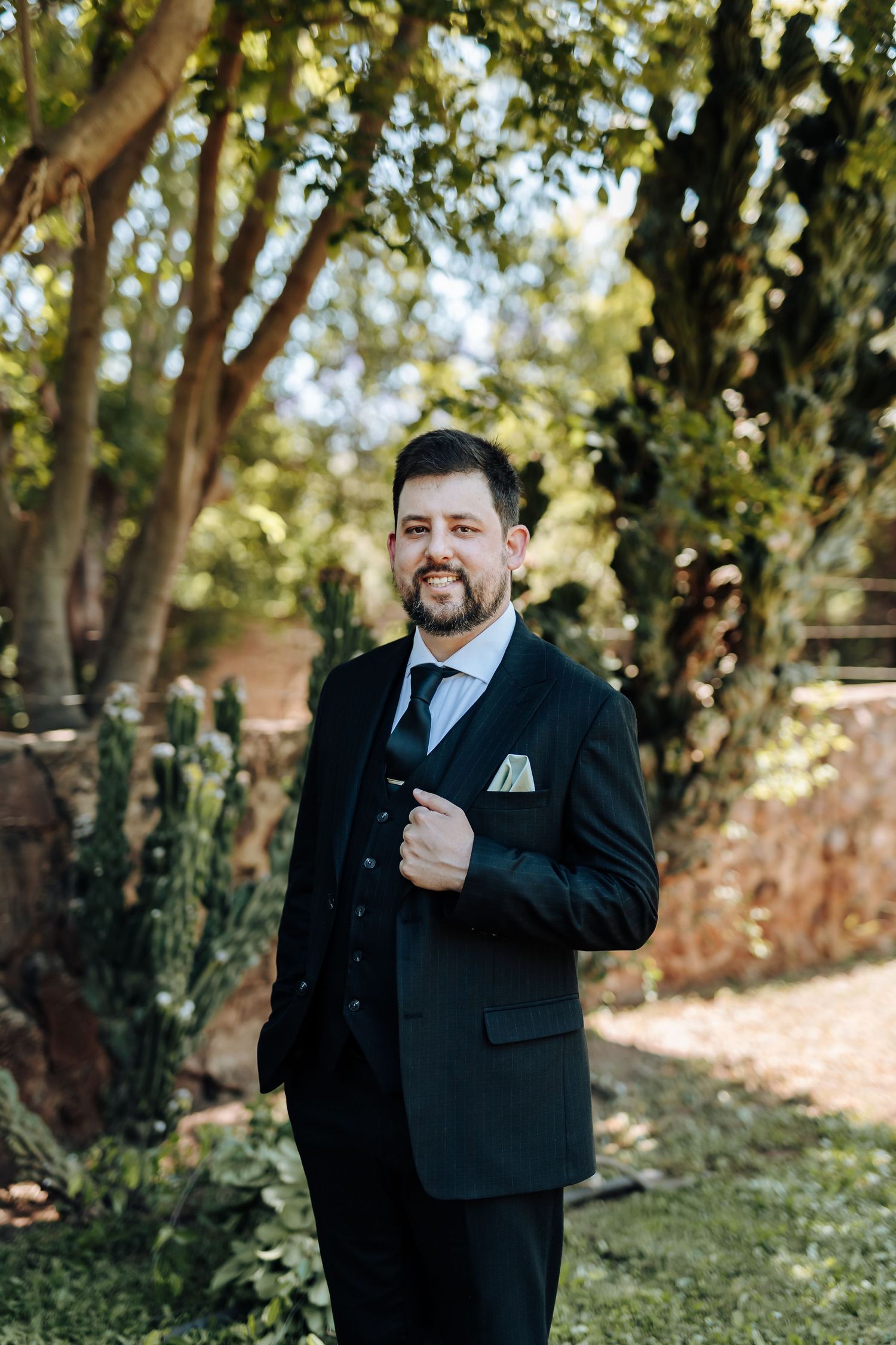 Wedding party members in black suits share a lighthearted moment outdoors among trees and greenery.