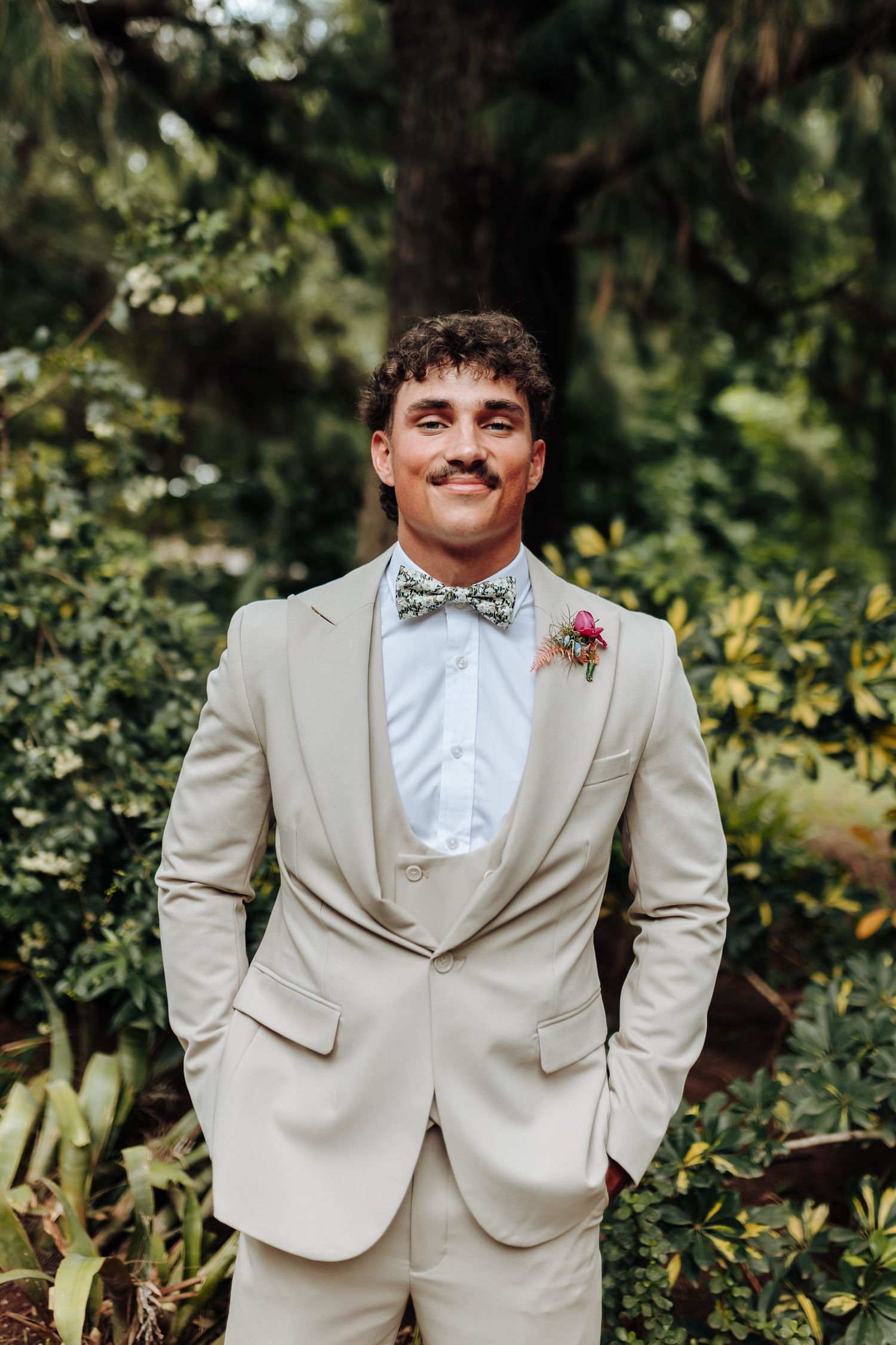 A groom in a light beige suit and bow tie smiles while standing in a lush garden setting.