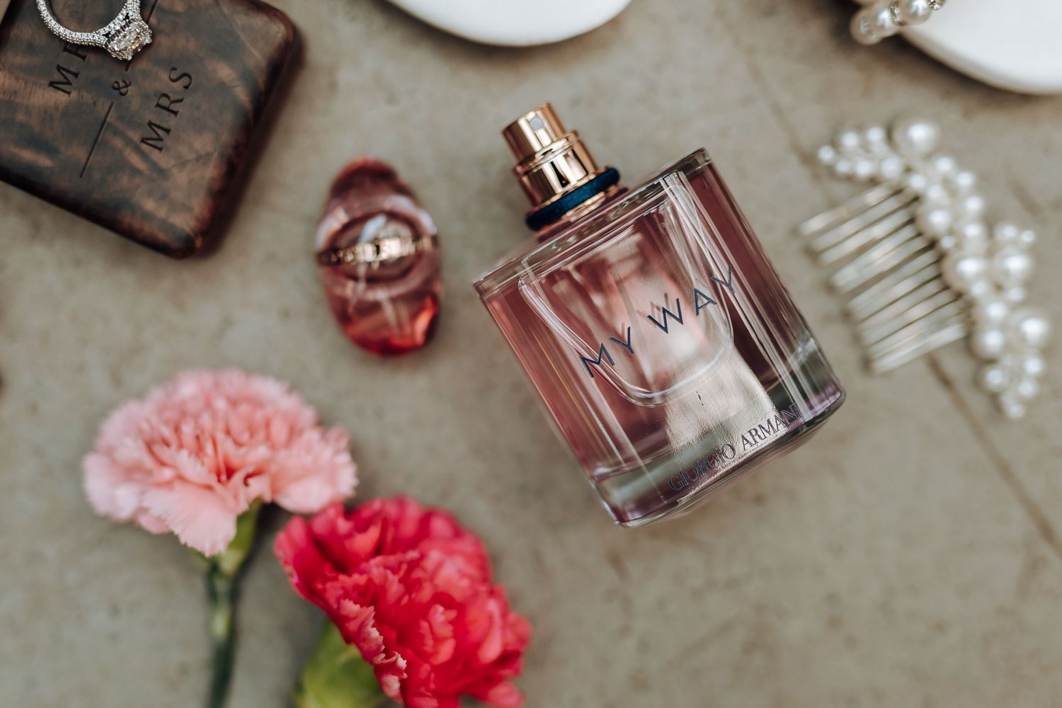 Pink and red carnations arranged with perfume bottles and delicate pearl accessories on a textured gray surface.
