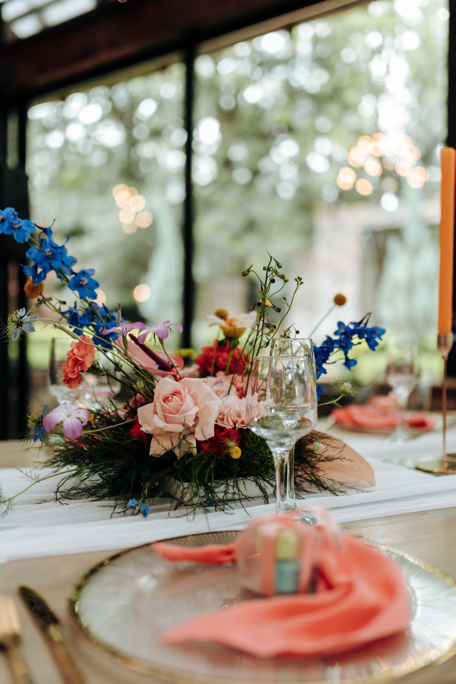 Elegant table setting with pink roses and blue delphinium centerpiece, coral napkin, and wine glass in soft natural light.