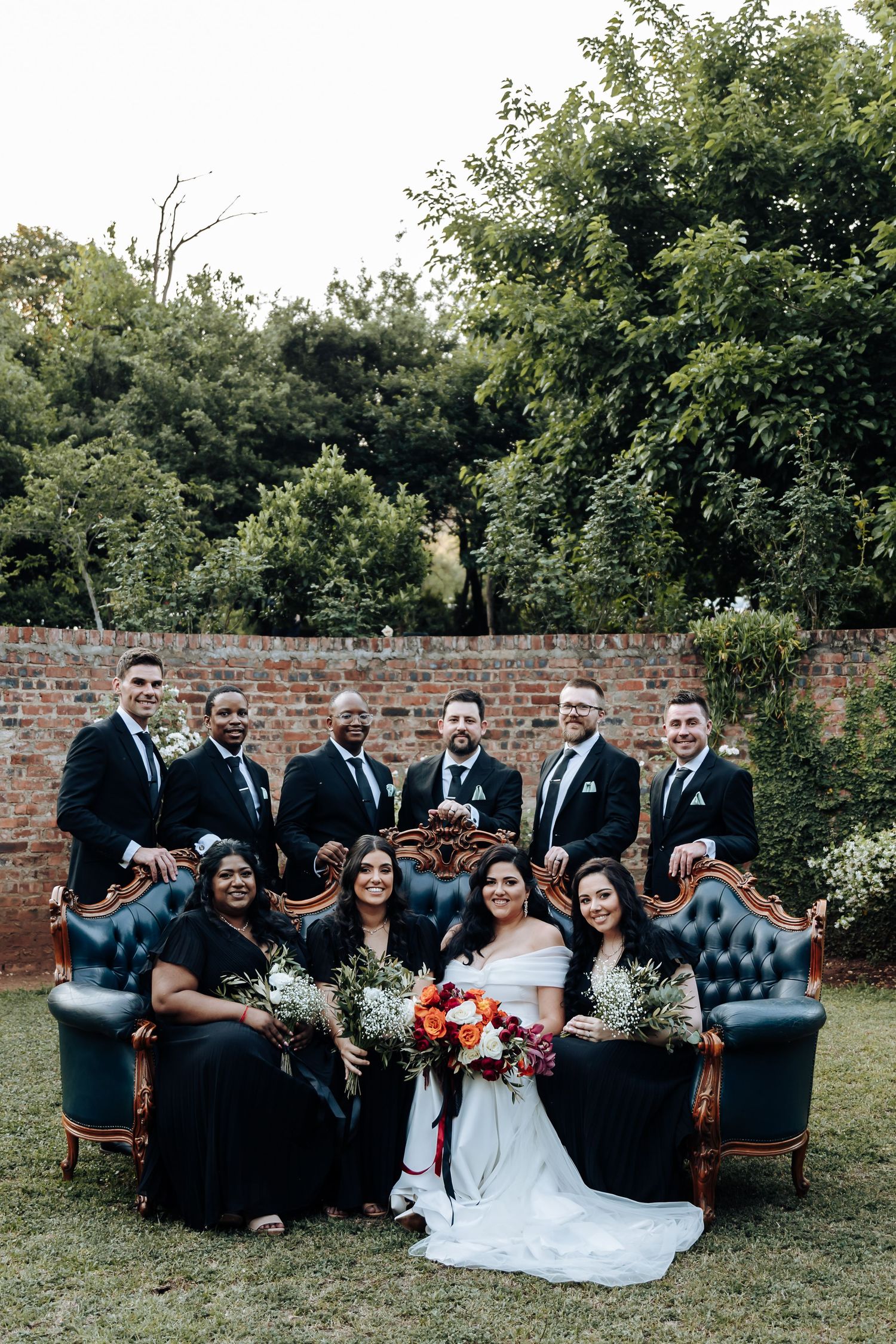 Wedding party poses on vintage blue couch in garden setting with brick wall backdrop.