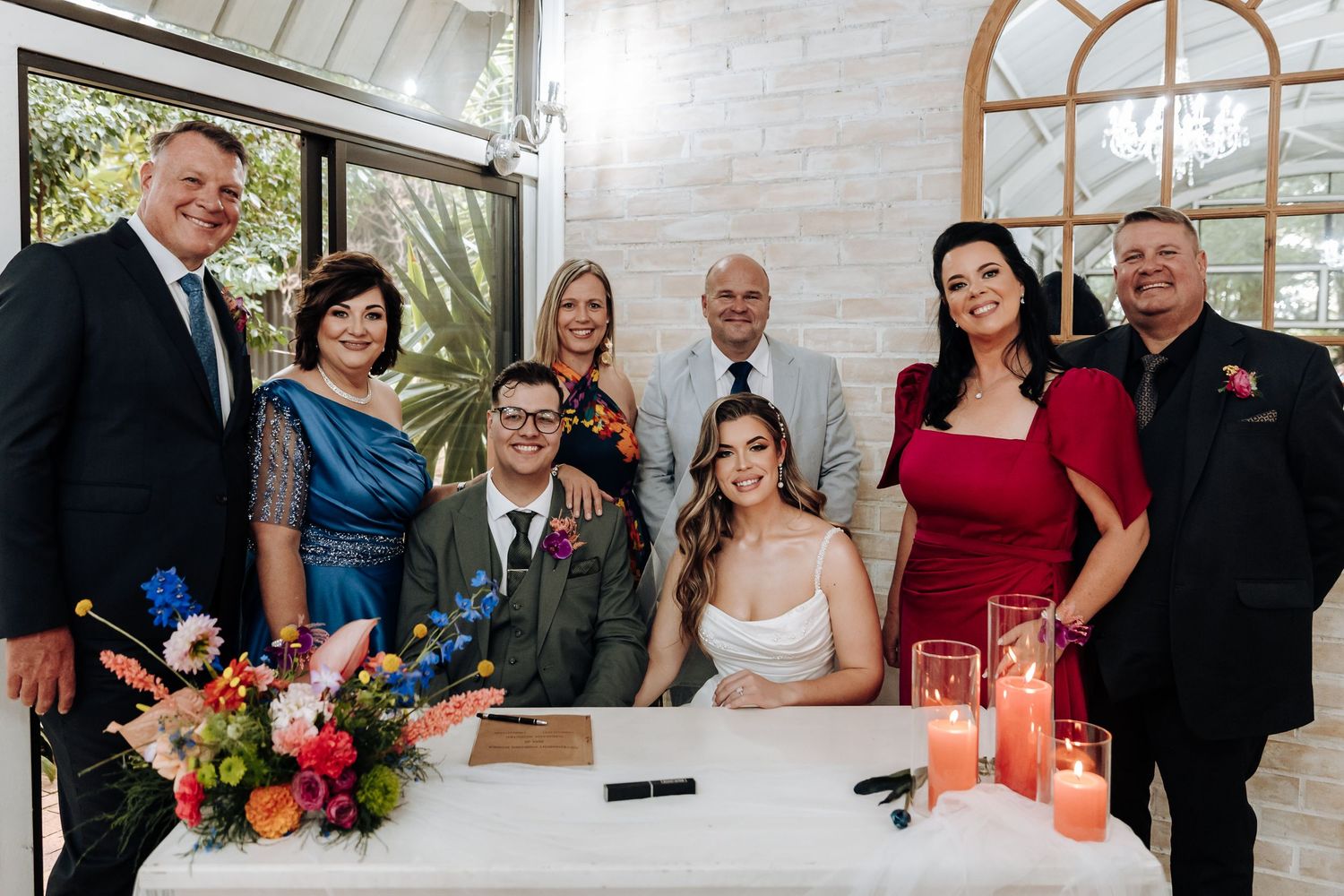 Wedding party gathers around signing table with floral arrangements and red candles in elegant venue.