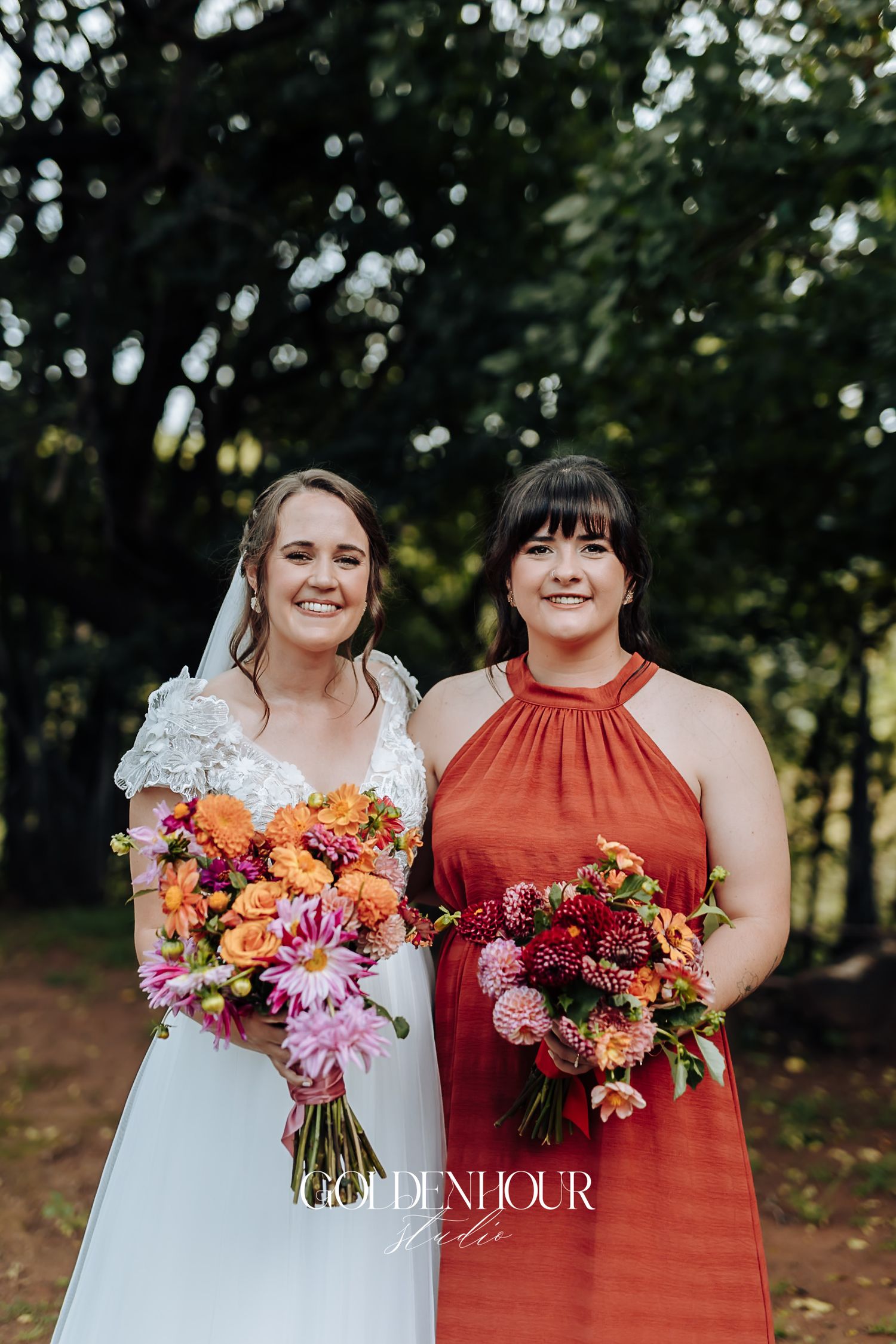 Bride in lace gown and bridesmaid in rust halter dress holding vibrant orange and pink bouquets.