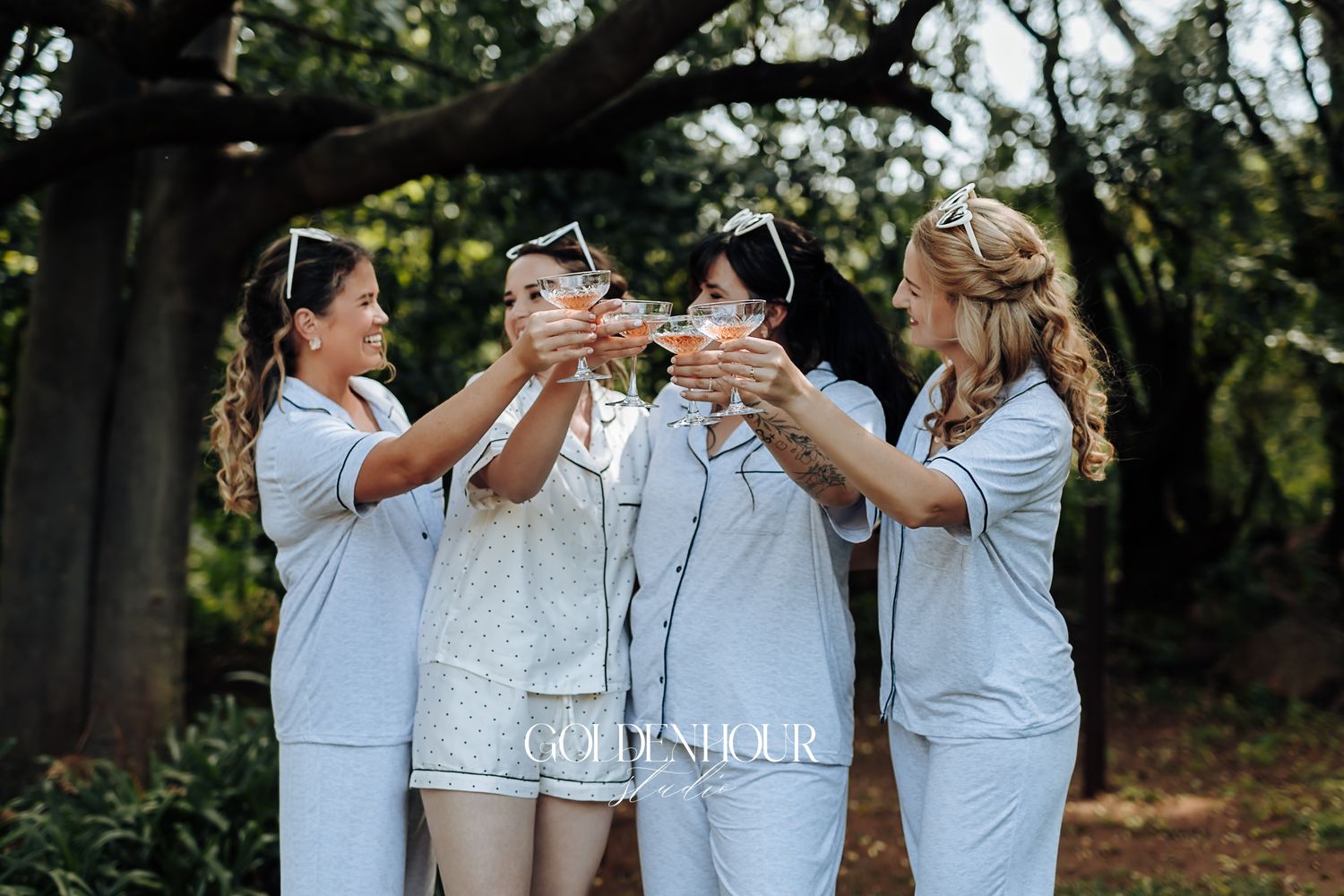 Four bridesmaids in matching blue pajamas toasting with champagne glasses outdoors under lush green trees.