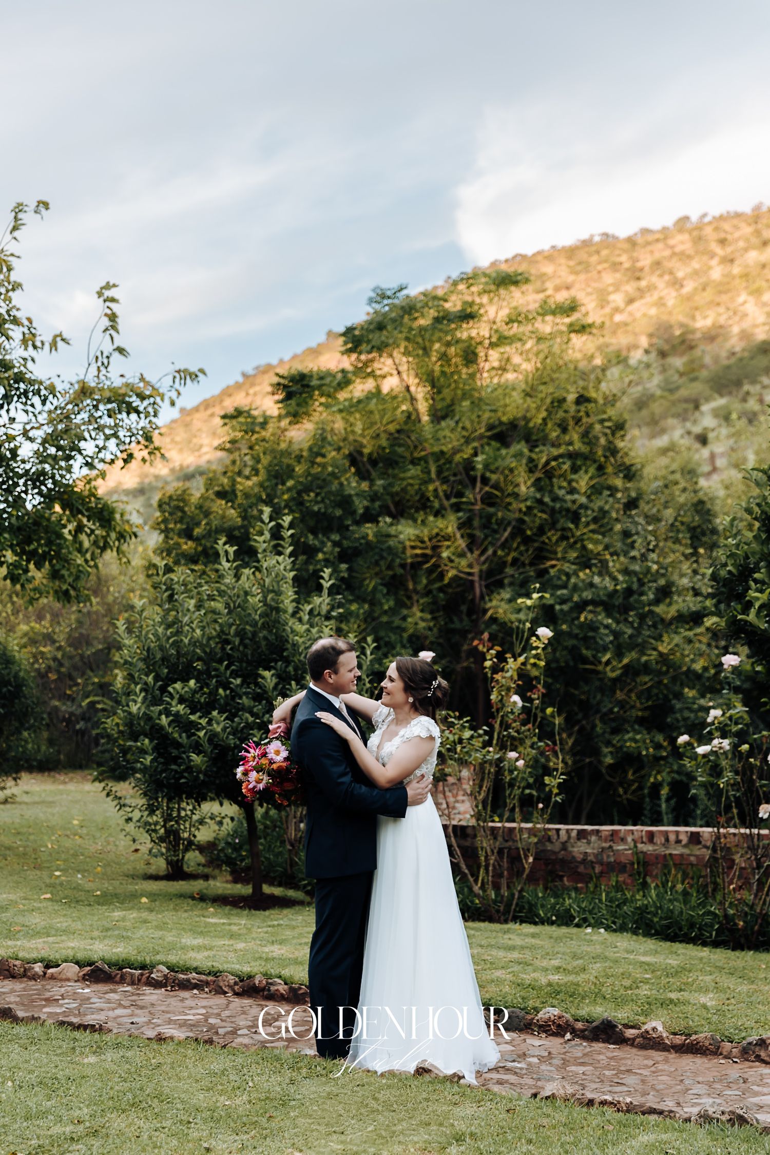 Bride and groom embrace romantically in lush garden with scenic mountain backdrop at sunset.