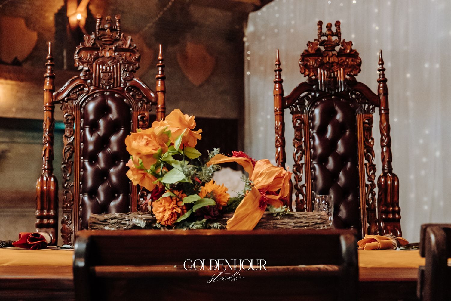 Two ornate dark wood throne chairs with tufted leather backs flank an orange floral centerpiece on a decorated sweetheart table.