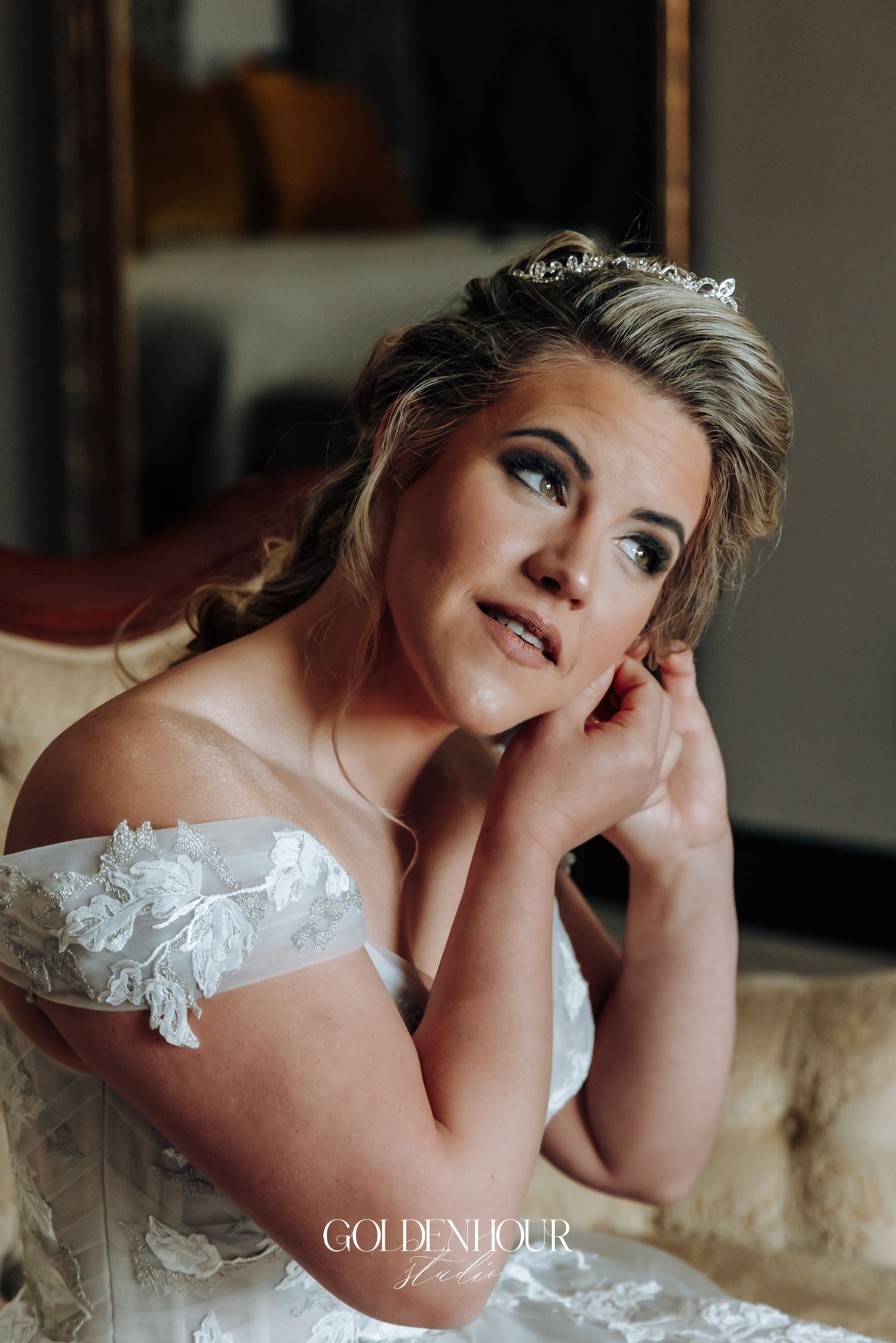 Beautiful bride in an off-shoulder lace wedding dress and tiara posing thoughtfully on an ornate vintage sofa.