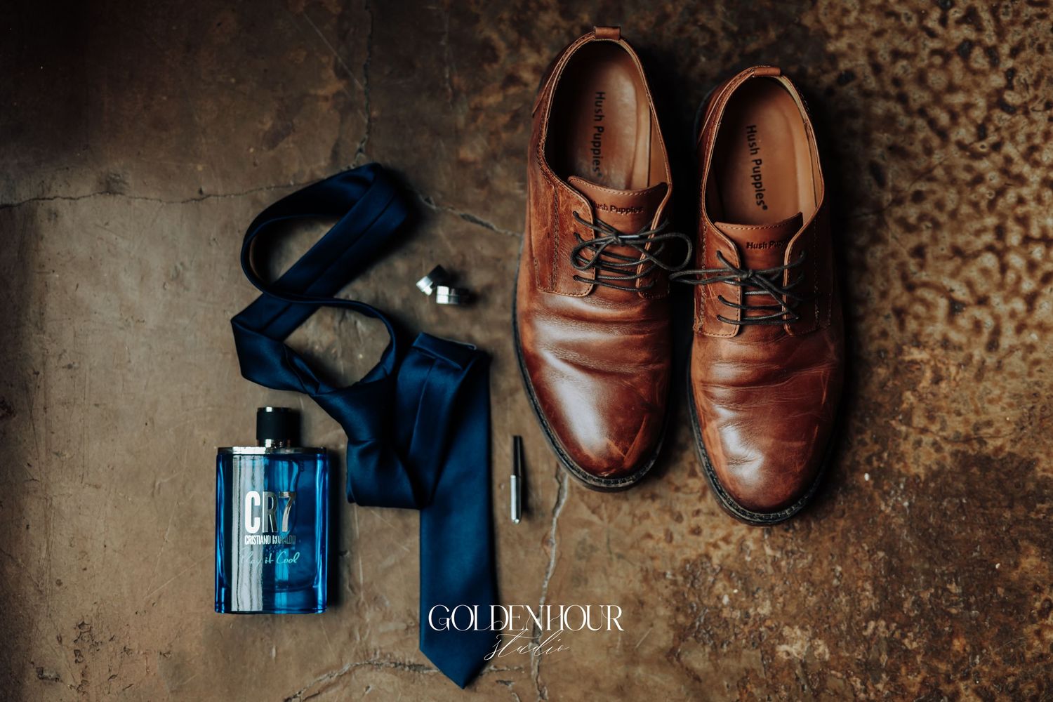 Brown leather dress shoes, blue silk tie, cufflinks, and blue cologne bottle arranged on rustic floor for wedding day.
