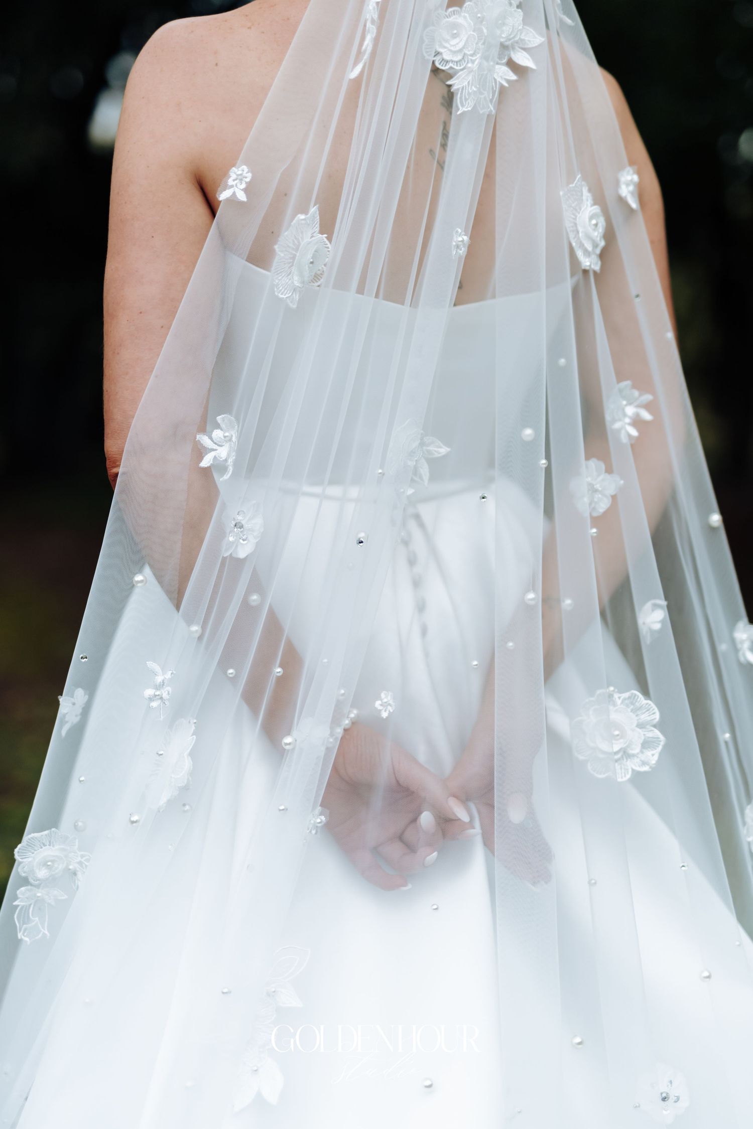 Bride in white wedding gown with floral embroidered veil adorned with pearls, viewed from behind.