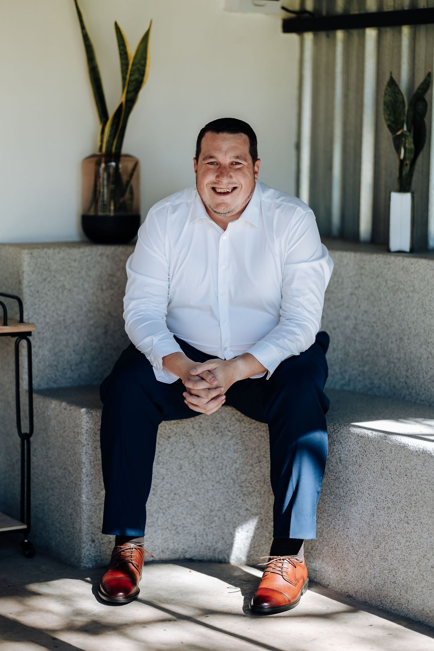 Smiling man in white shirt and navy pants with brown dress shoes sitting on stone steps with plants in background.