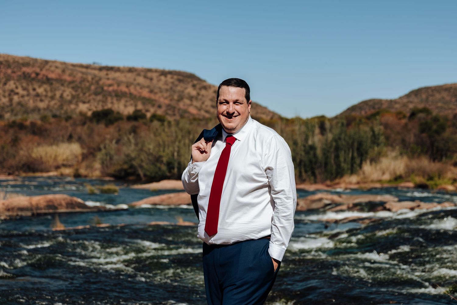 Man in white shirt and red tie smiling by a scenic river with rocky hills in the background.