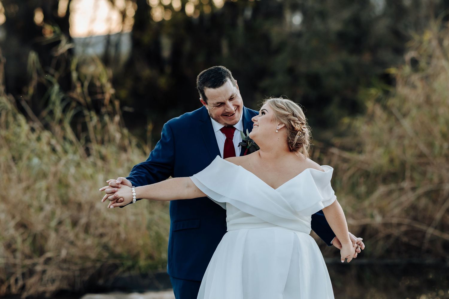 Bride in white off-shoulder gown and groom in navy suit dance outdoors near trees in a romantic wedding photo.