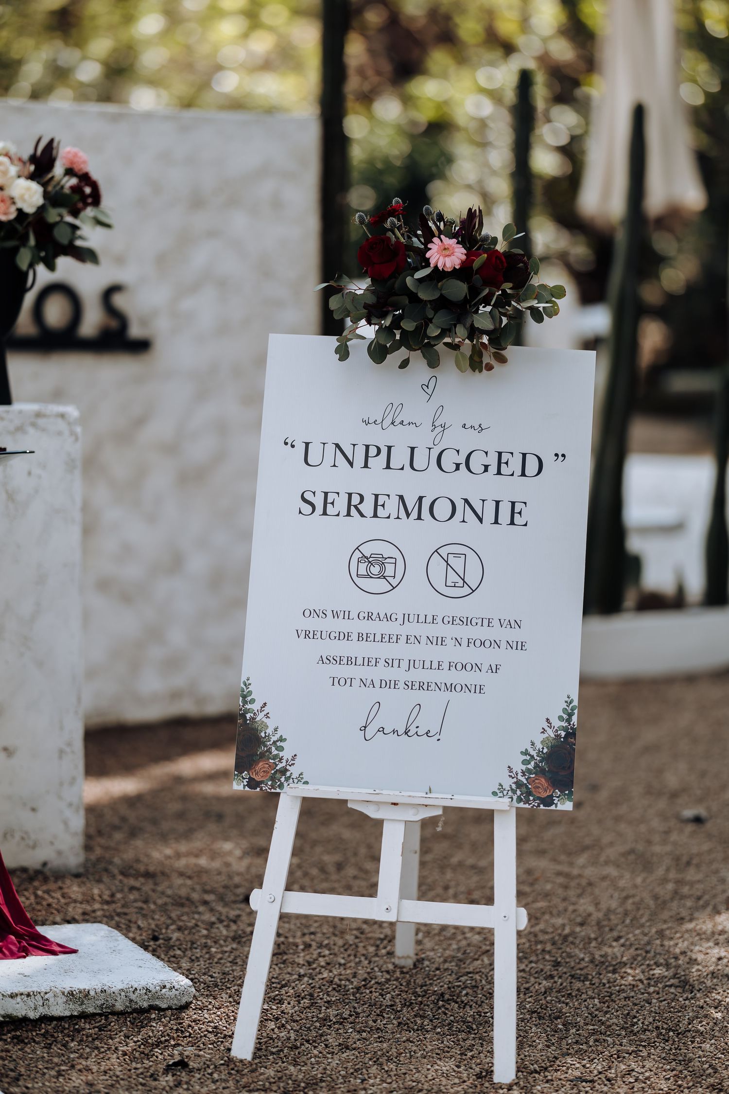 White easel sign for an unplugged wedding ceremony decorated with greenery and burgundy flowers.