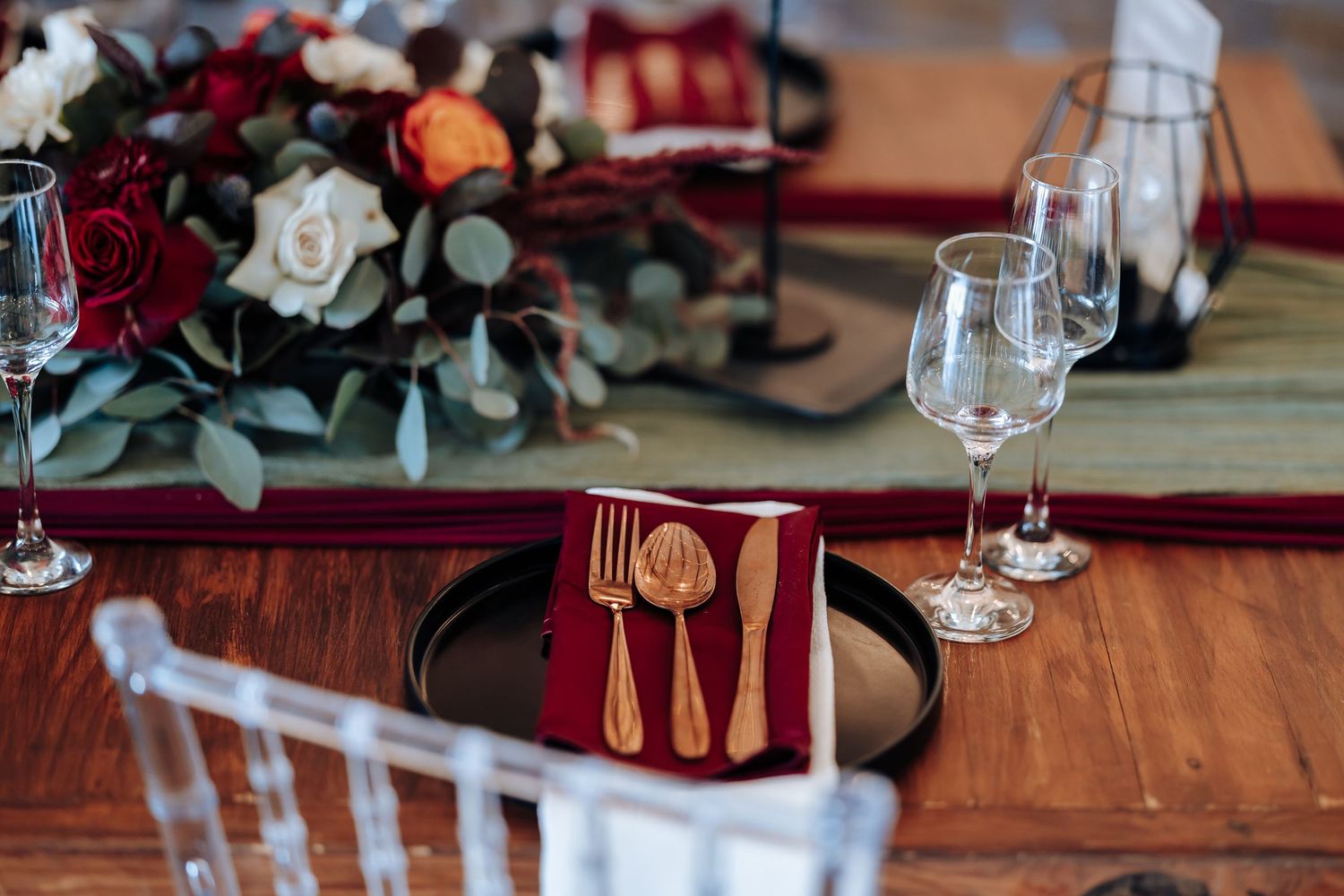 Elegant wedding table setting with gold cutlery on red napkin, crystal glasses, and floral centerpiece with red and white roses.