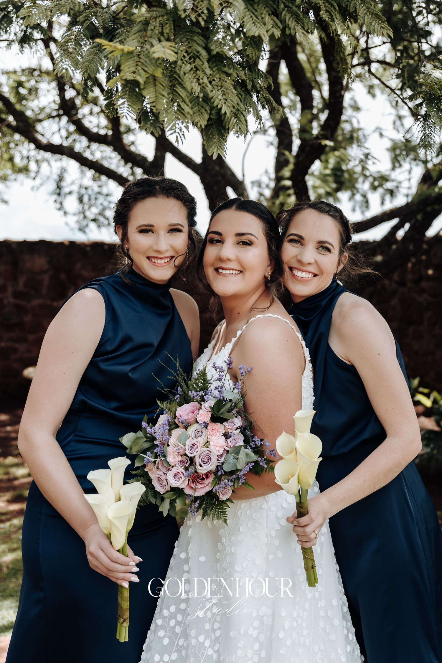 Wedding party poses under tree with bridesmaids in navy dresses holding calla lilies.