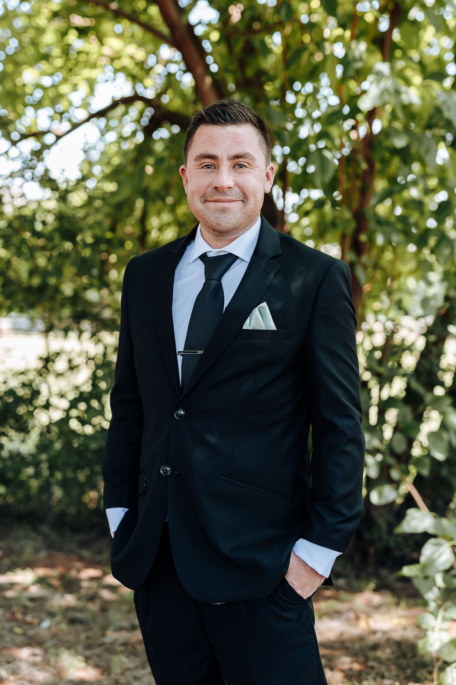 A person in a black suit and tie poses outdoors with trees and natural lighting in the background.