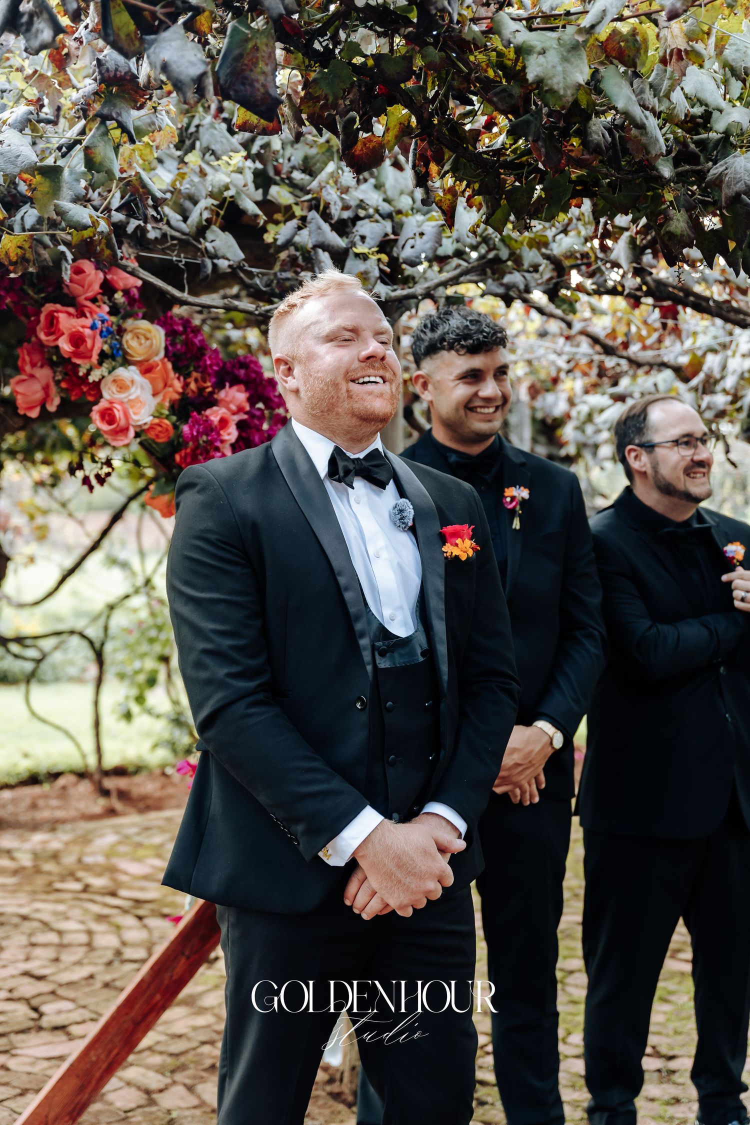 Groomsmen in black suits share laughter and candid moments under flowering trees during an outdoor wedding celebration.