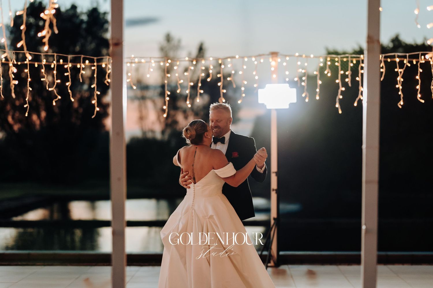A romantic wedding couple dances under twinkling string lights at dusk during their outdoor reception celebration.