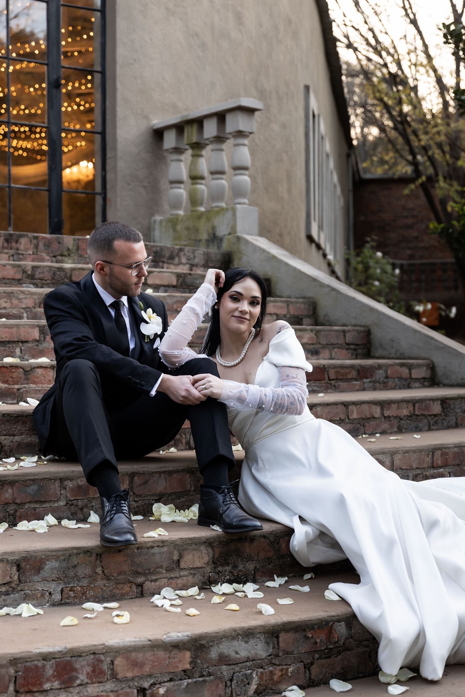 A couple in wedding attire sits together on brick steps with flower petals scattered around at dusk.