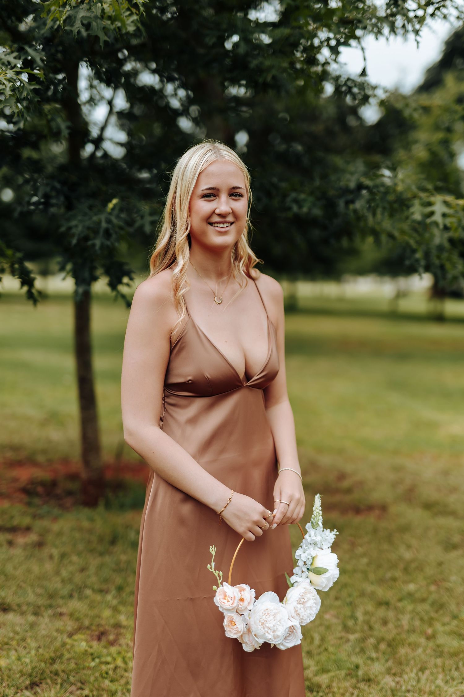 A person in a brown satin dress holds a white floral bouquet while standing in a grassy outdoor setting.