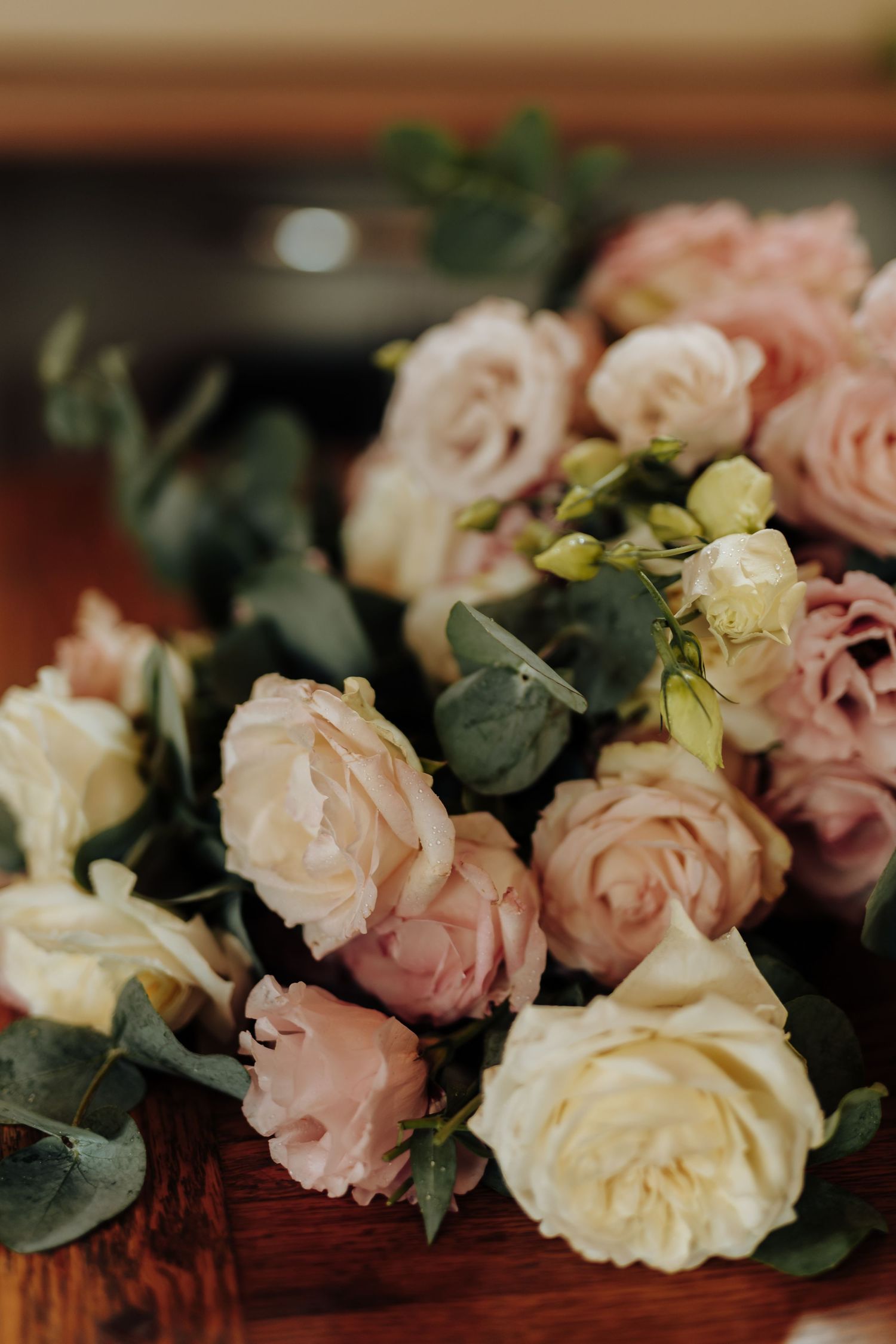 Beautiful pink and cream roses with eucalyptus greenery arranged on a rich wooden surface in soft lighting.