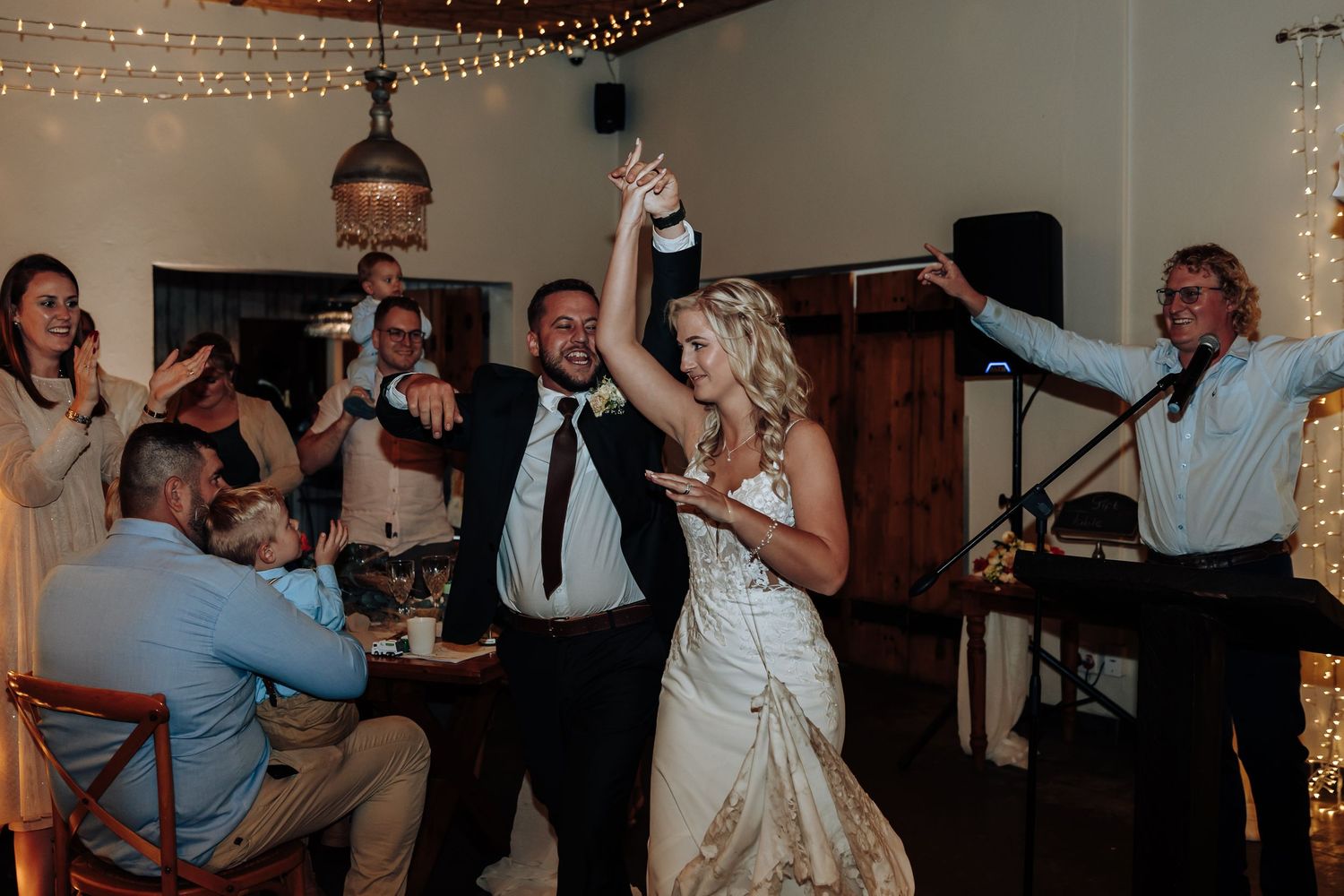 People dancing and celebrating at a wedding reception under string lights and wooden beams.