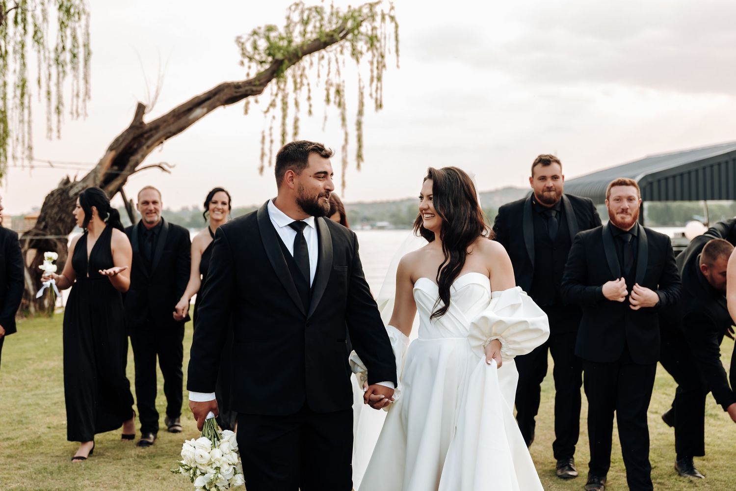 Wedding party in formal attire poses together under a willow tree during outdoor celebration.