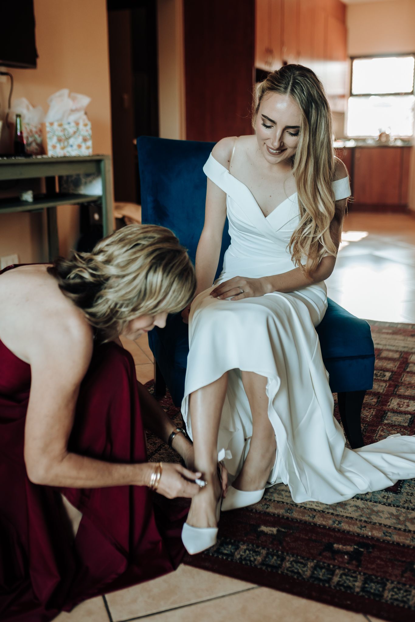 A wedding preparation moment as someone helps adjust a white shoe on a bride seated in a blue chair.