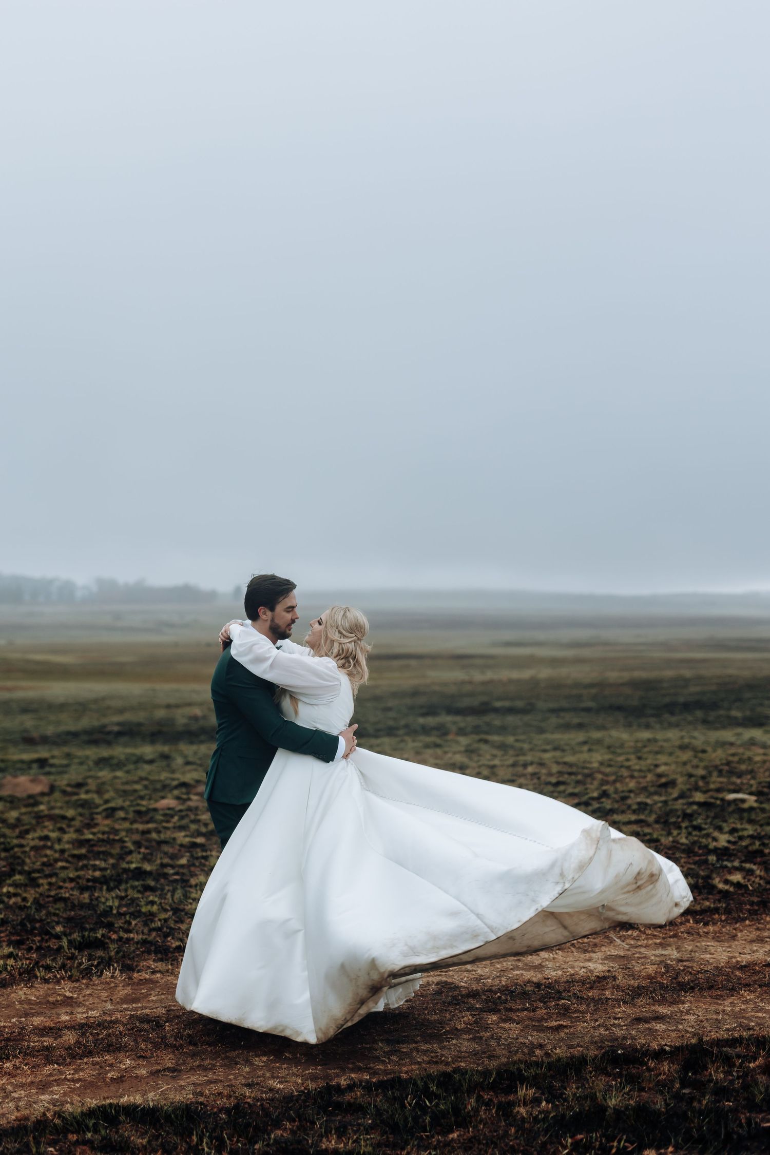 Dancing newlyweds embrace on a dirt path as the bride's dress billows in the wind.