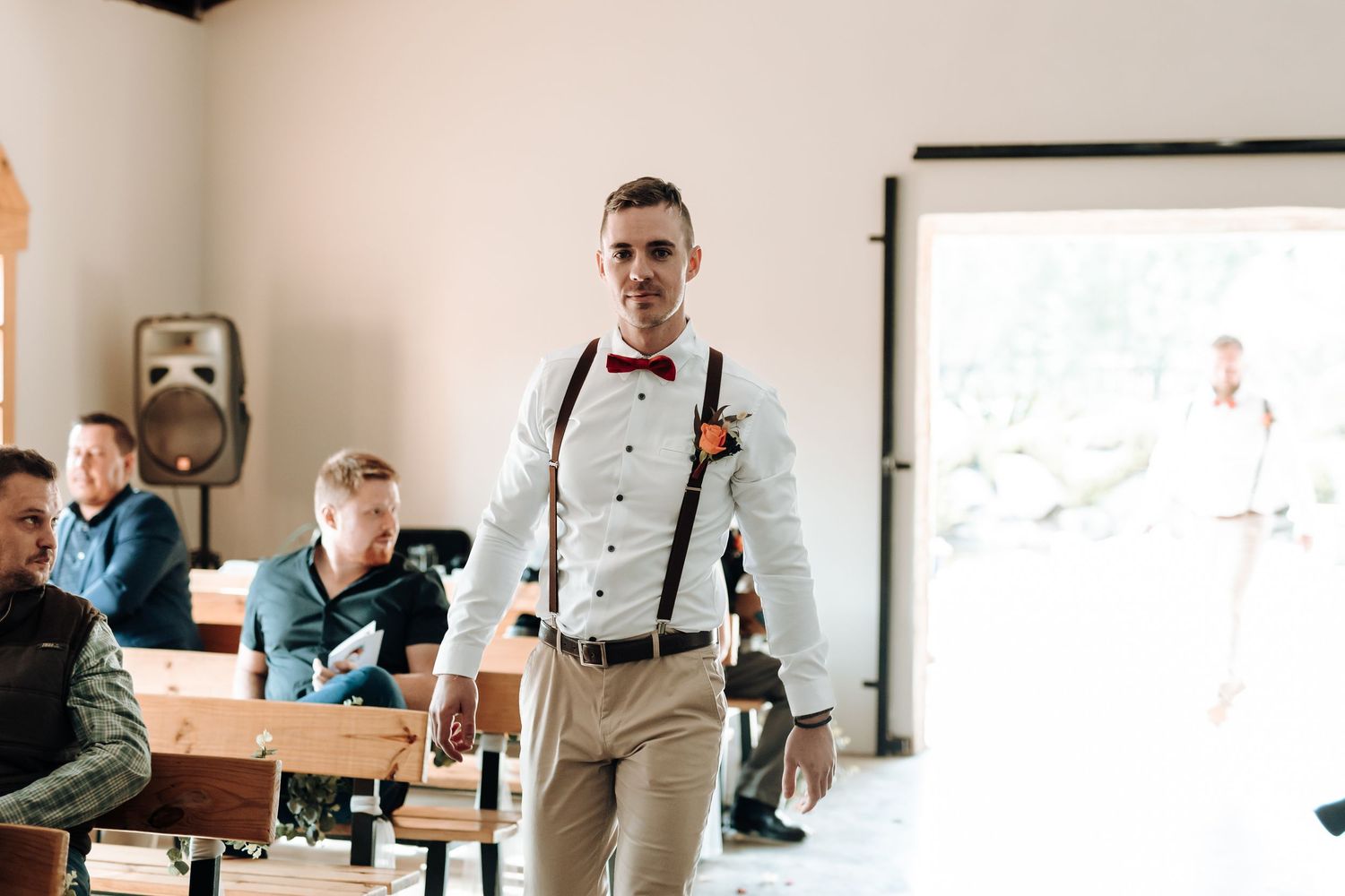 Stylish groom in white shirt and suspenders enters venue with natural lighting.