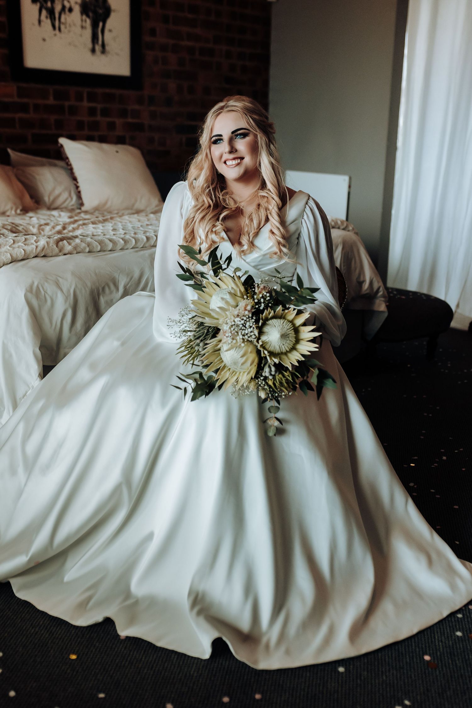 Bride seated in elegant white dress holding dried flower bouquet against brick wall backdrop.