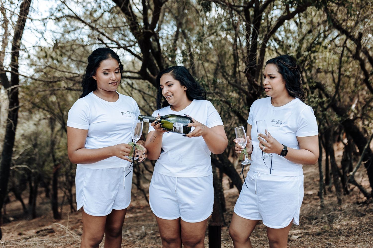 Three people in matching white outfits sharing drinks outdoors among trees.