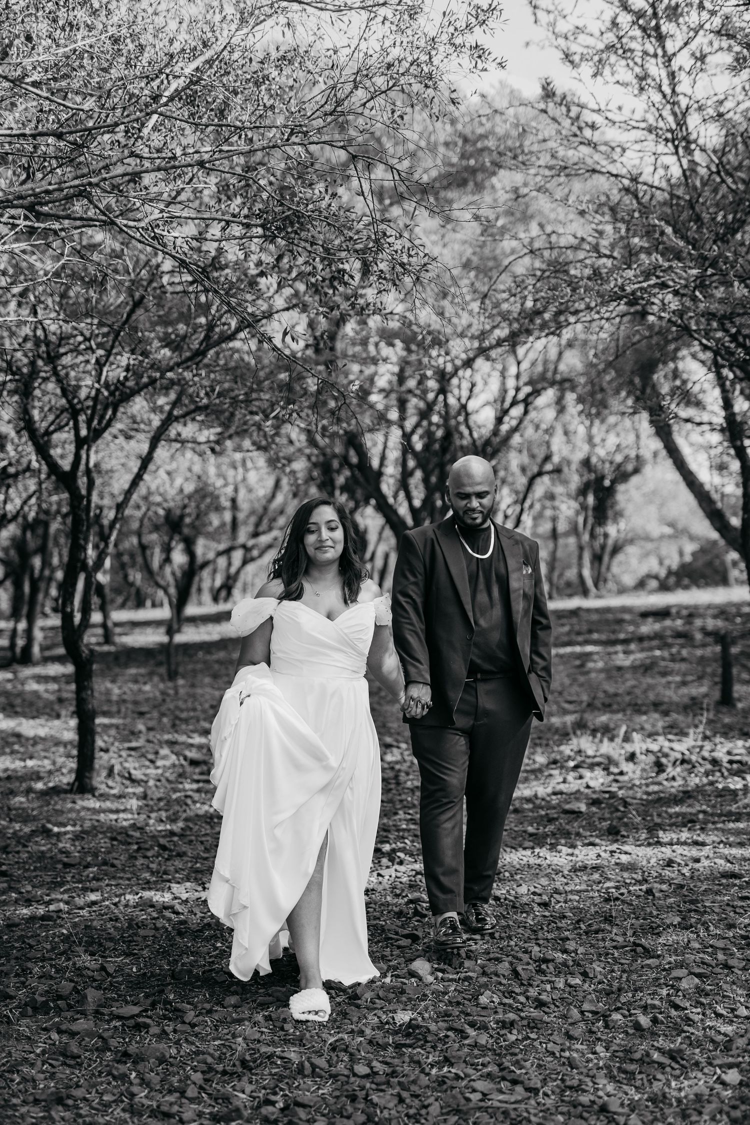 Black and white photo of bride and groom walking through tree-lined path.
