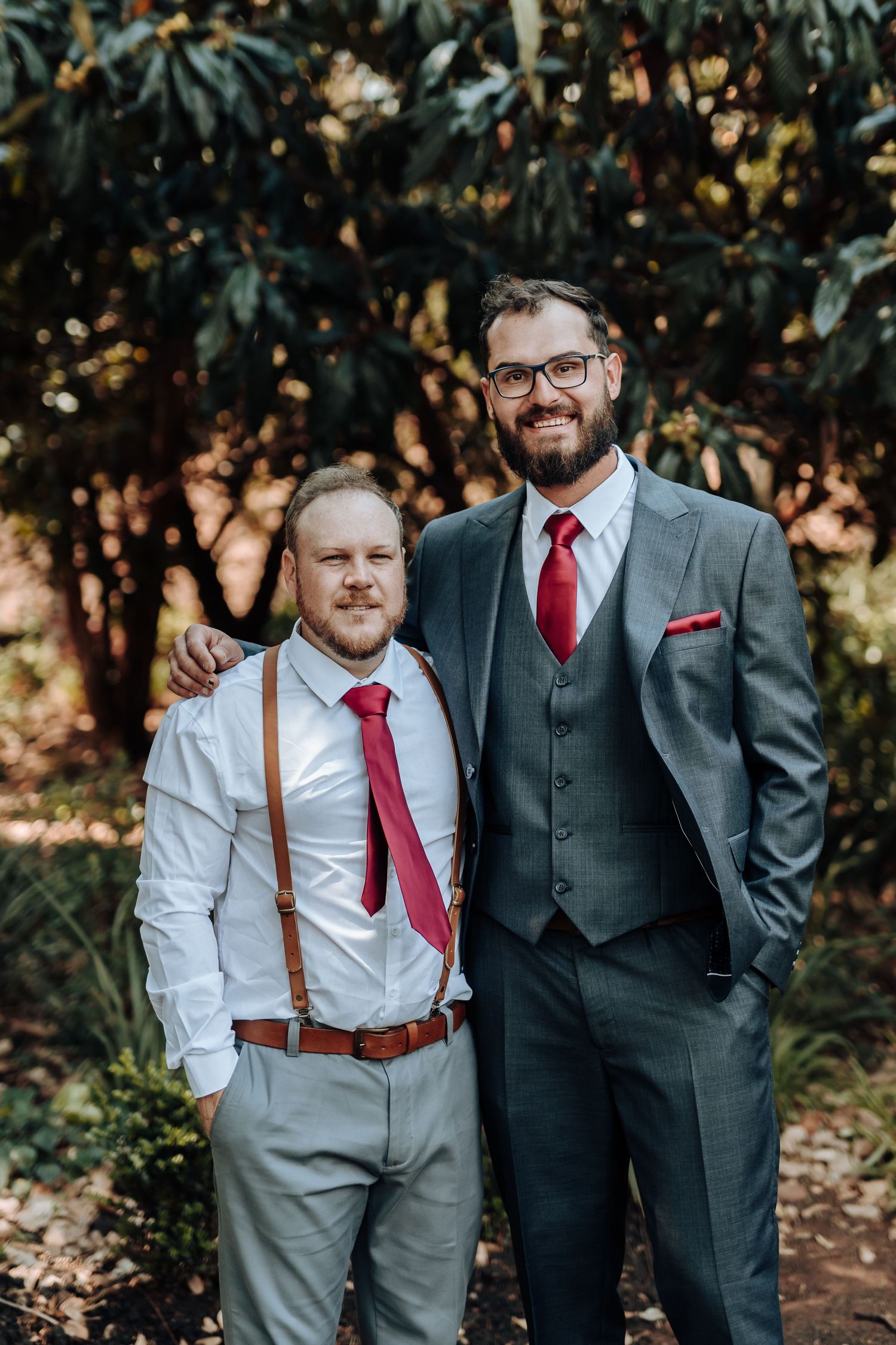 Wedding party members in contrasting suits pose together in outdoor garden setting.