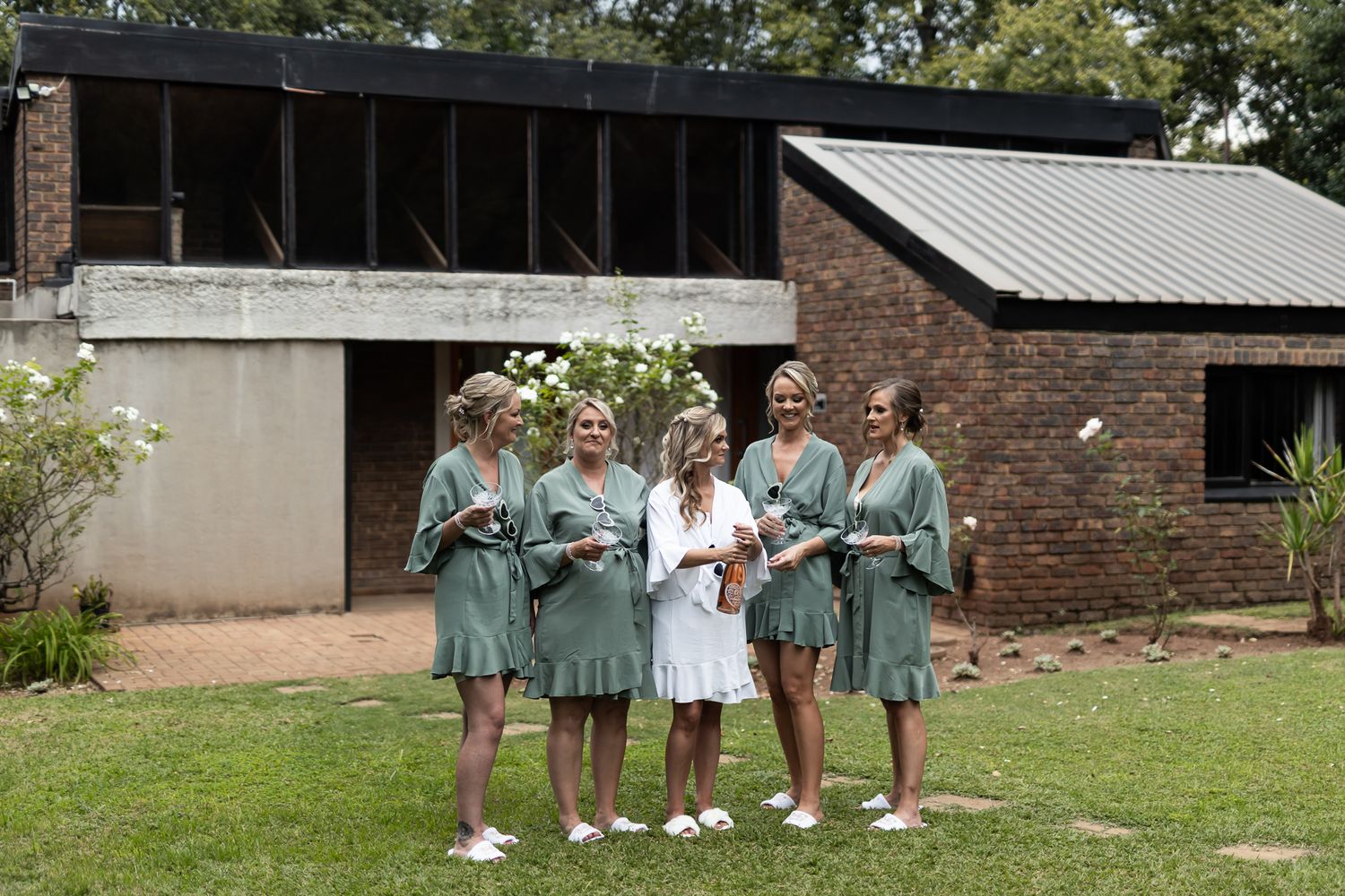 Bridesmaids in sage green robes pose with the bride in white on a grassy lawn.