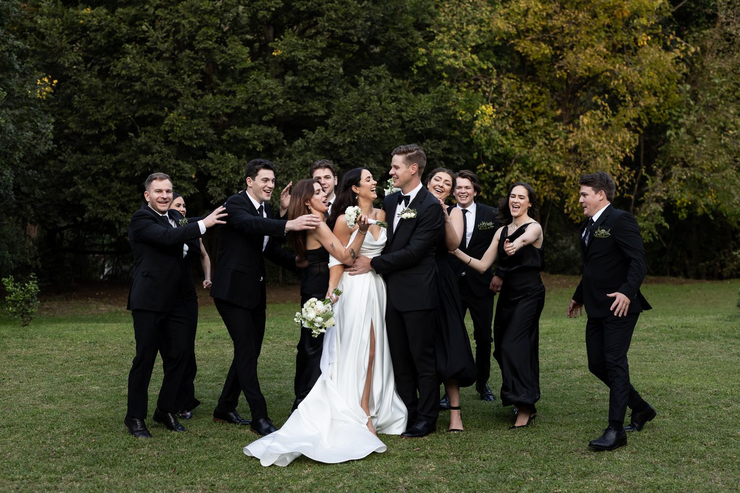 A wedding party poses casually on grass while sharing laughs together.