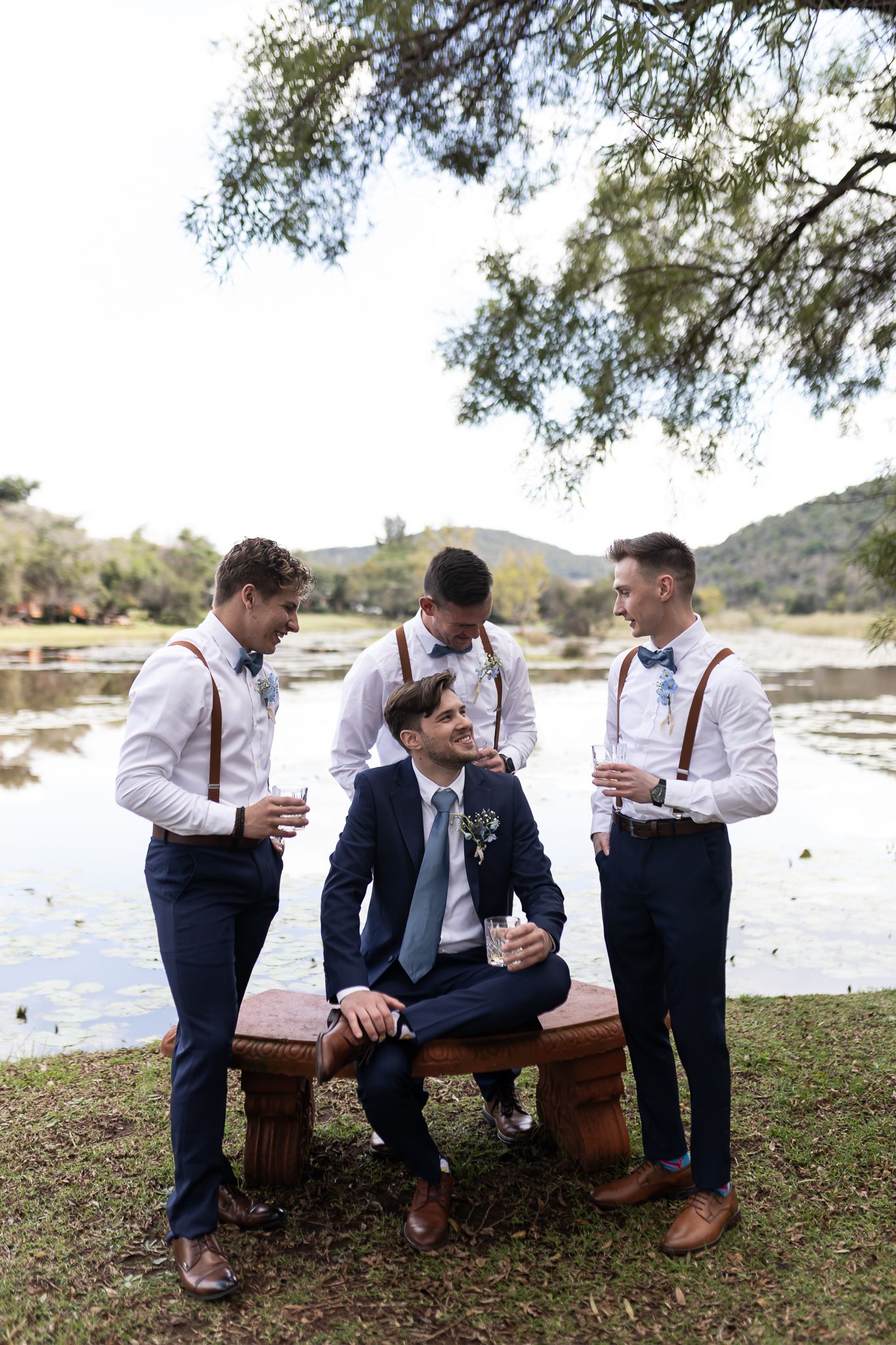 Wedding party members in formal attire gather around a bench near a lake.