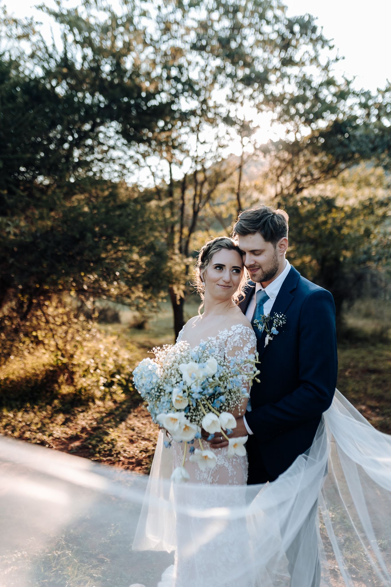 Elegant wedding couple poses on a sunlit path with autumn trees and flowing white veil in background.