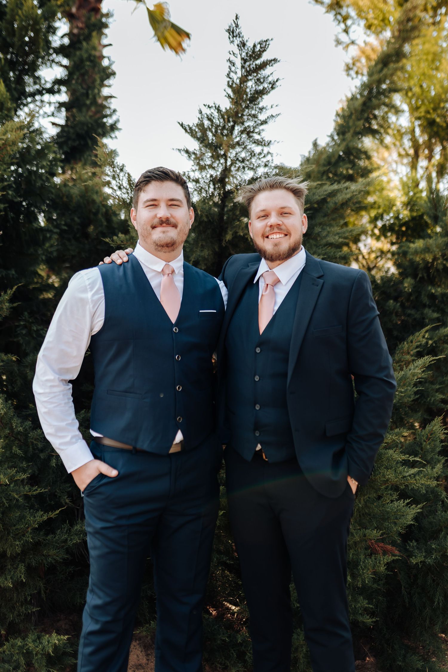 Two men in navy suits with pink ties stand together outdoors among evergreen trees during a formal event.