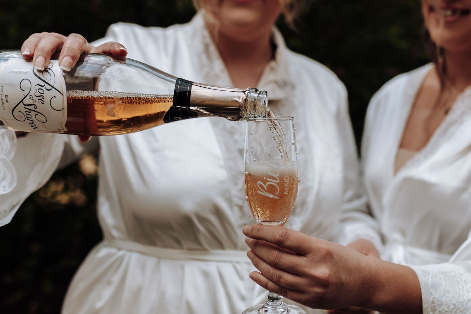 Person wearing white robe pours champagne into a delicate glass flute during celebratory preparations.