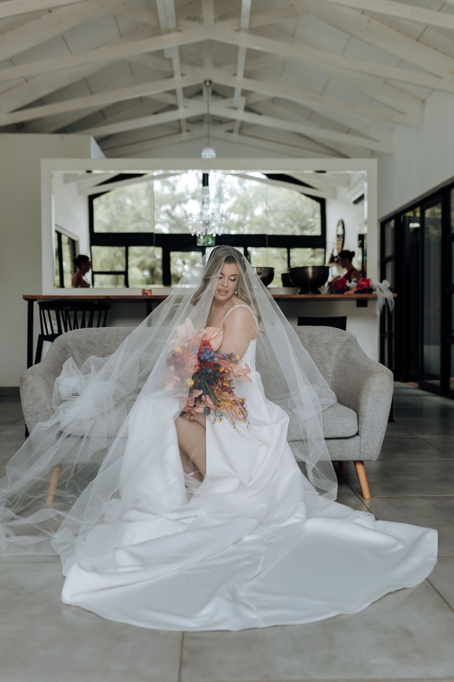 A flowing white wedding gown spreads across a light-colored floor beneath crystal chandeliers.
